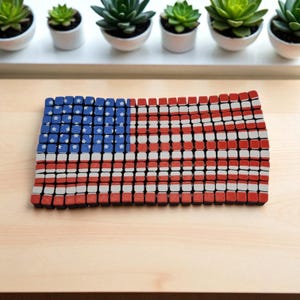 Può includere: Una bandiera americana a mosaico fatta di piccoli blocchi quadrati. La bandiera presenta un campo blu con stelle bianche nell'angolo in alto a sinistra e strisce rosse e bianche. È esposta su una superficie di legno chiaro.