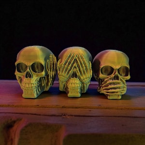 May include: Three yellow-green skulls, each with a different hand gesture. One skull has hands over its ears, another over its eyes, and the third over its mouth. The skulls are on a wooden surface with a dark background.