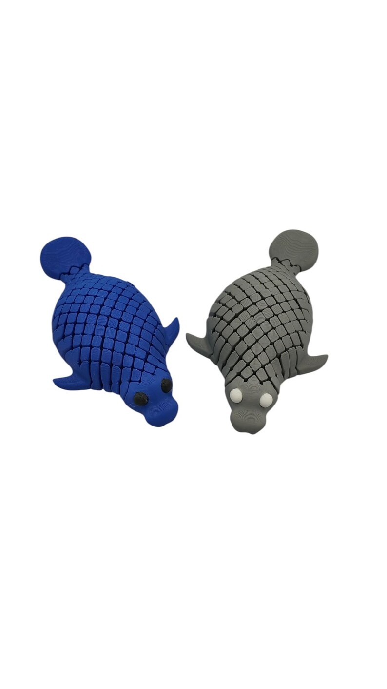 Puede incluir: Dos objetos con forma de foca. Uno es azul, el otro gris. Ambos tienen un patr&oacute;n texturizado en forma de escamas en sus cuerpos, con peque&ntilde;as aletas y una cabeza redondeada. La foca azul tiene ojos negros, la foca gris tiene ojos blancos.