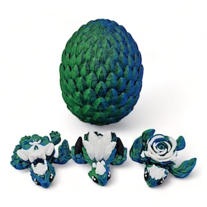 Puede incluir: Tres peque&ntilde;as figuras de drag&oacute;n verde y azul con detalles blancos. Un drag&oacute;n tiene una rosa blanca en la espalda, otro tiene una flor blanca y el tercero tiene un ala blanca. Los dragones est&aacute;n sentados sobre una superficie blanca. Un huevo de drag&oacute;n verde y azul m&aacute;s grande est&aacute; en el fondo.
