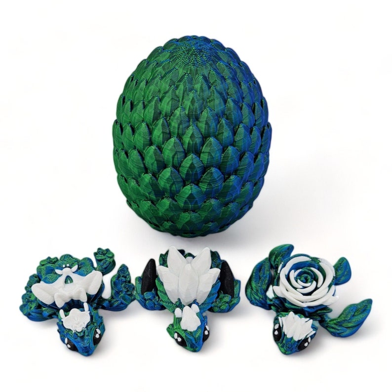 Puede incluir: Tres peque&ntilde;as figuras de drag&oacute;n verde y azul con detalles blancos. Un drag&oacute;n tiene una rosa blanca en la espalda, otro tiene una flor blanca y el tercero tiene un ala blanca. Los dragones est&aacute;n sentados sobre una superficie blanca. Un huevo de drag&oacute;n verde y azul m&aacute;s grande est&aacute; en el fondo.
