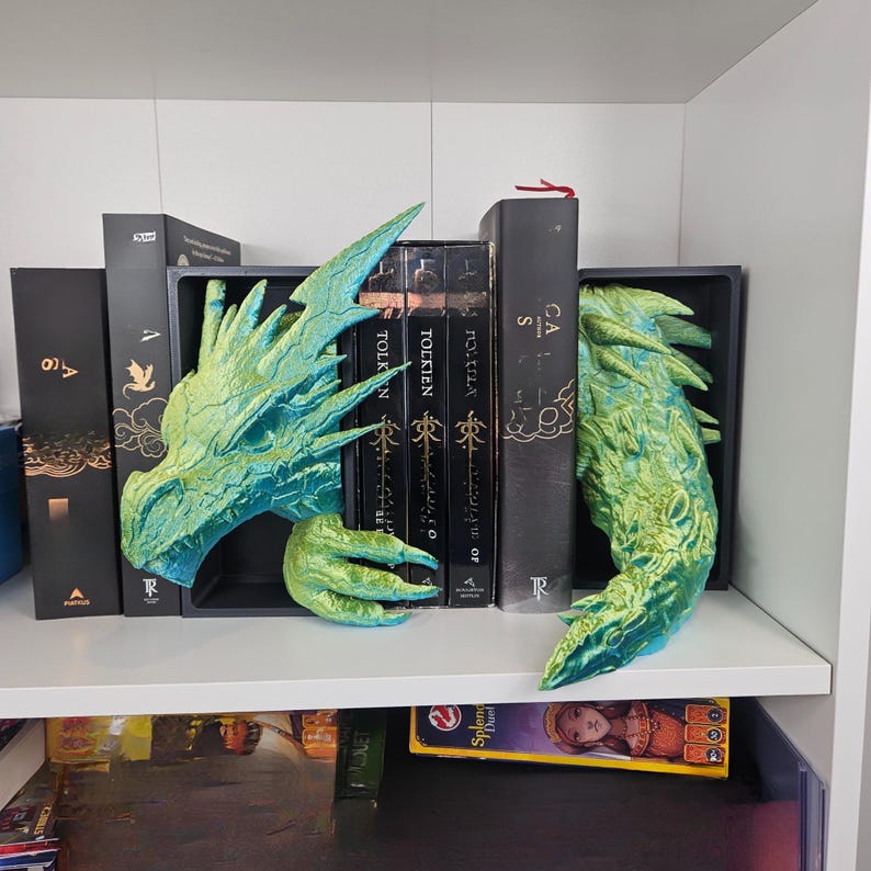 May include: A pair of dragon-themed bookends in shades of green and blue. The bookends are placed on a white shelf, holding up a collection of books. The dragon's head and tail are visible, adding a fantasy touch to the bookshelf.