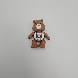 May include: A brown bear figurine with a white belly featuring a coffee cup design. The bear has a smiling face, small black eyes, and brown arms and legs. The figurine is set against a plain white background.