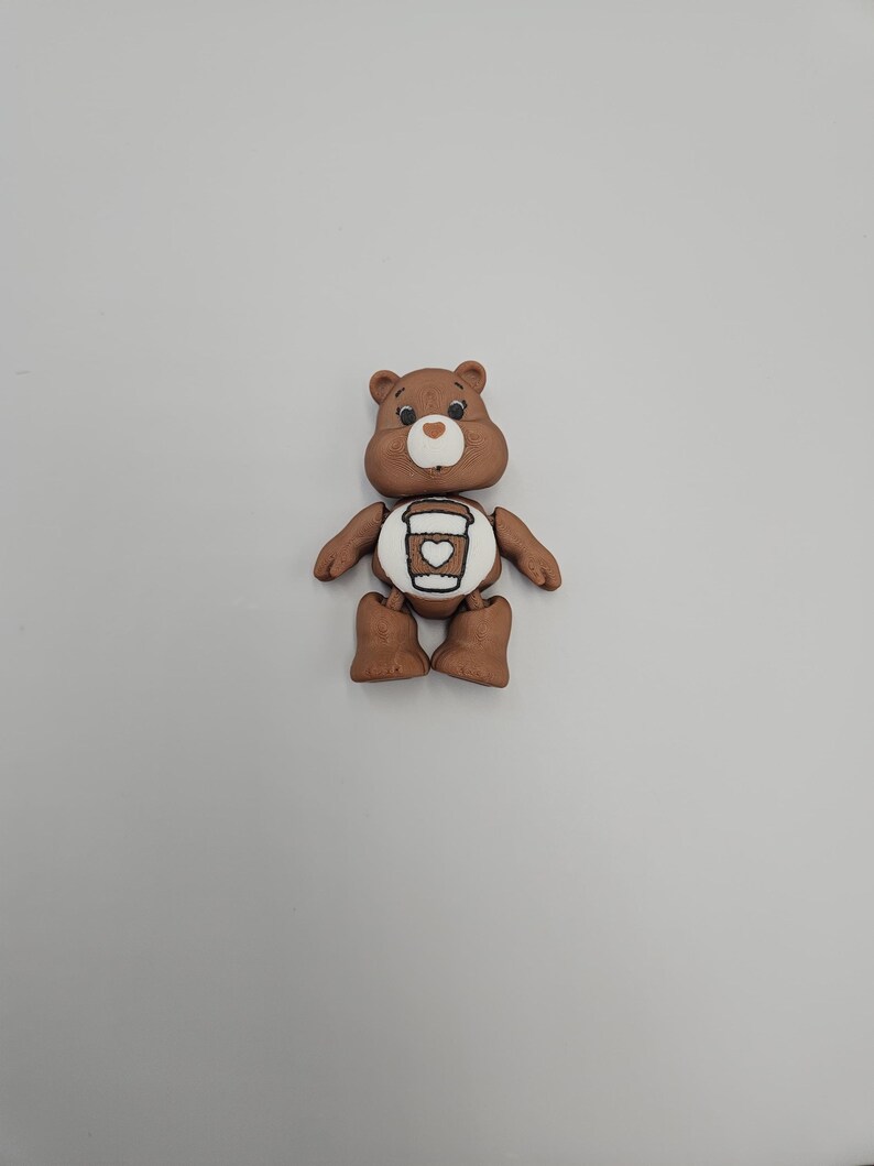 May include: A brown bear figurine with a white belly featuring a coffee cup design. The bear has a smiling face, small black eyes, and brown arms and legs. The figurine is set against a plain white background.