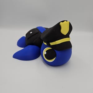Pode incluir: Um personagem de desenho animado azul, preto e amarelo usando um chap&eacute;u de policial. O personagem tem um corpo azul grande e redondo, p&eacute;s azuis e uma parte traseira preta. O chap&eacute;u de policial &eacute; preto com uma faixa e um distintivo amarelos.