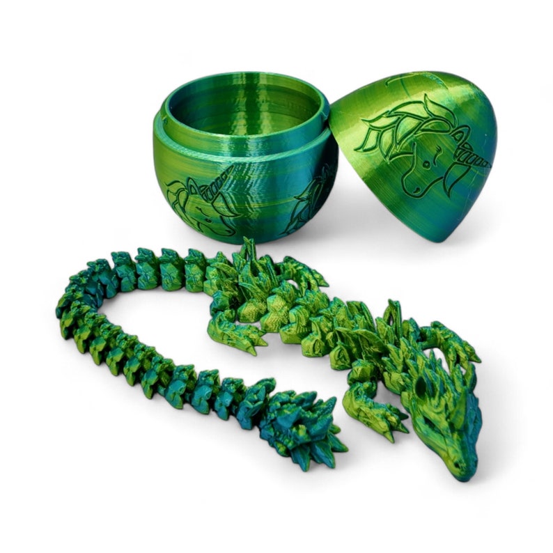May include: A green and blue 3D printed dragon figurine with a segmented body. The dragon is curled up in a circle. The dragon is in a closed egg-shaped container with a unicorn design. The container is also green and blue.