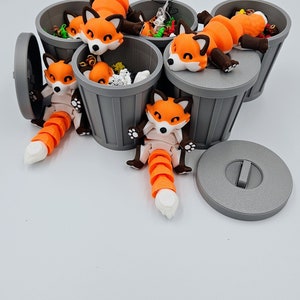 May include: A collection of orange and white fox figurines lounging on and in small gray trash cans. The foxes have articulated tails and are posed in various positions. The trash cans are filled with small, colorful objects.