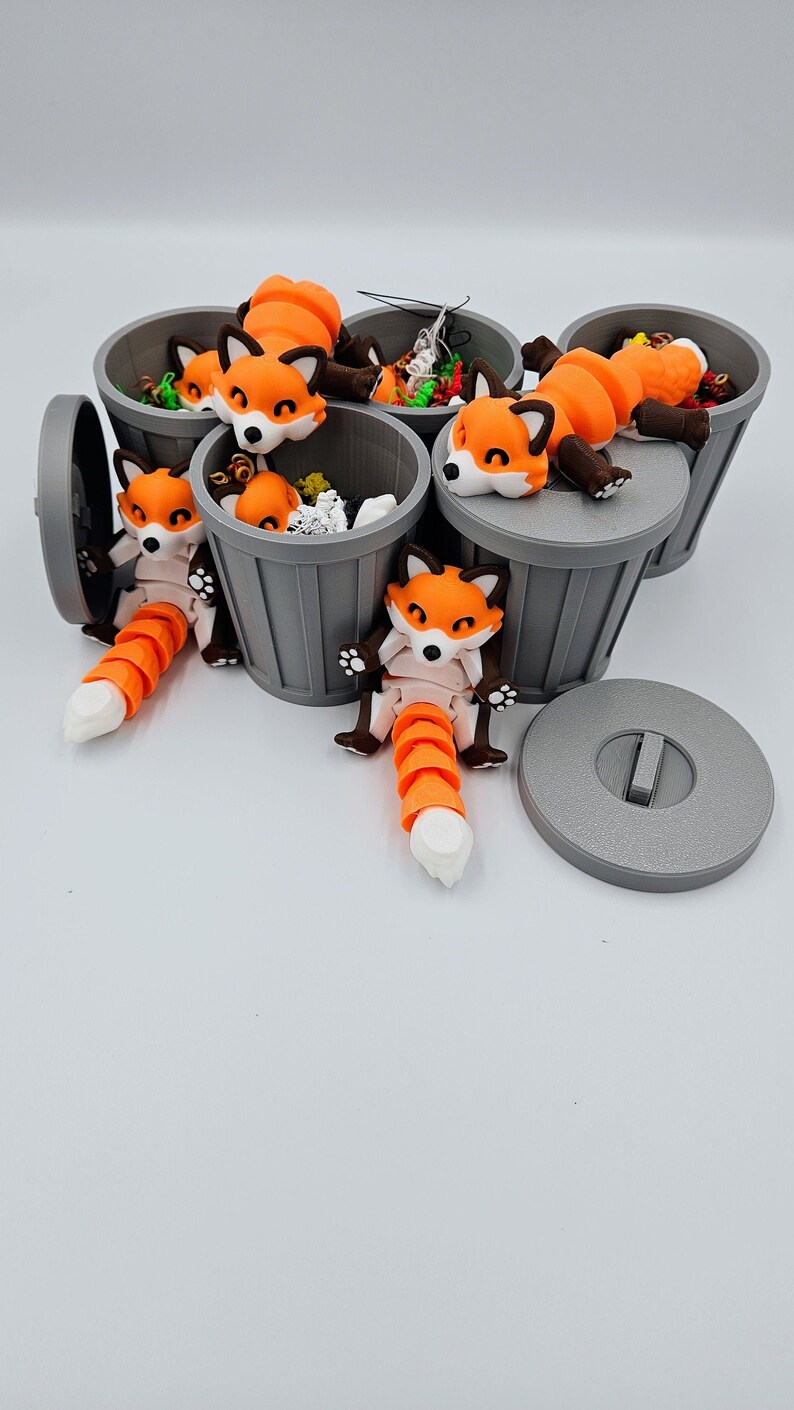 May include: A collection of orange and white fox figurines lounging on and in small gray trash cans. The foxes have articulated tails and are posed in various positions. The trash cans are filled with small, colorful objects.