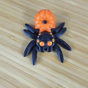 Puede incluir: Un juguete de ara&ntilde;a de pl&aacute;stico negro y naranja con un cuerpo en forma de calabaza y ocho patas. La ara&ntilde;a tiene ojos grandes y redondos con pupilas amarillas.