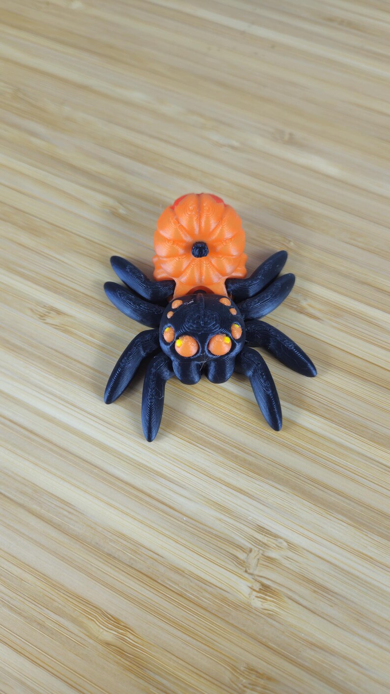Puede incluir: Un juguete de ara&ntilde;a de pl&aacute;stico negro y naranja con un cuerpo en forma de calabaza y ocho patas. La ara&ntilde;a tiene ojos grandes y redondos con pupilas amarillas.
