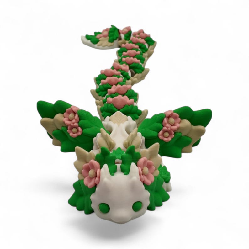May include: A whimsical, handcrafted figurine of a dragon, featuring a cream-colored head and body, adorned with green leaves and pink flowers. The dragon's wings and tail are also decorated with leaves and flowers, creating a fantasy-themed sculpture.