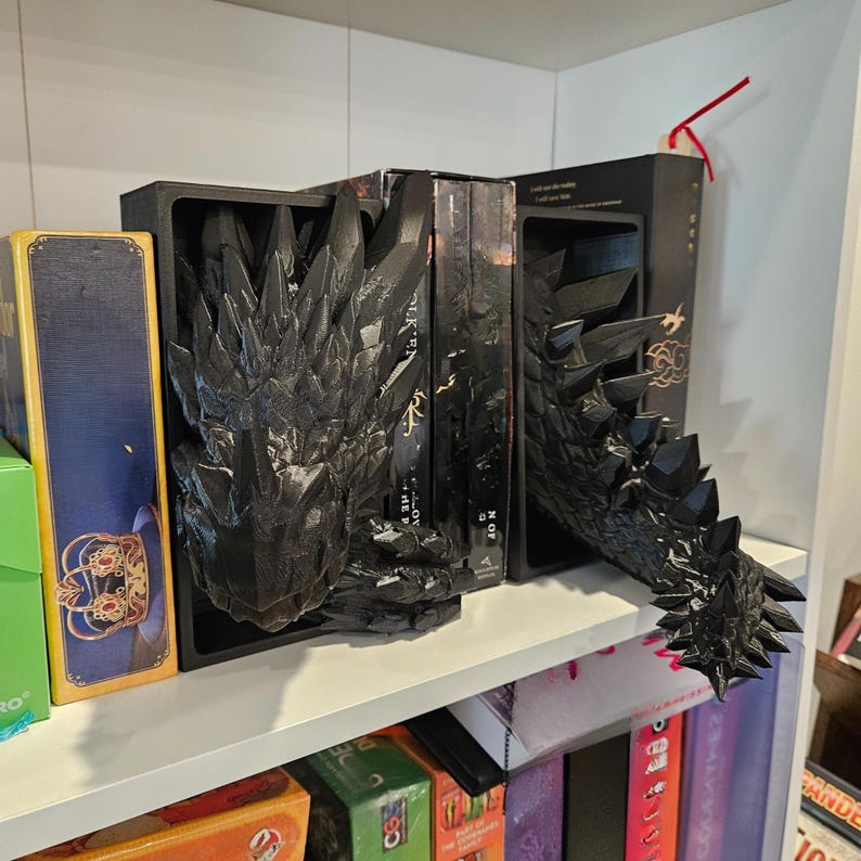 Op de afbeelding: Zwarte drakenboekensteunen met een gedetailleerd, gestructureerd ontwerp. De boekensteunen hebben een drakenkop en -staart, waardoor een display met fantasiethema ontstaat. De boekensteunen staan op een witte plank met boeken.