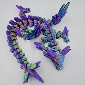 May include: A 3D printed dragon figurine with a purple, green, and blue iridescent finish. The dragon is posed in a curled position with its head raised and its wings spread out.