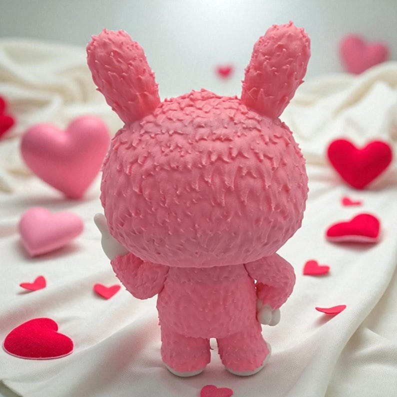 K&ouml;nnte beinhalten: Eine rosa, strukturierte Hasenfigur mit gro&szlig;en Ohren und rundem K&ouml;rper, die auf einer wei&szlig;en Oberfl&auml;che steht. Der Hase ist von rosa und roten herzf&ouml;rmigen Dekorationen umgeben, was ein Valentinstag-Thema ergibt.