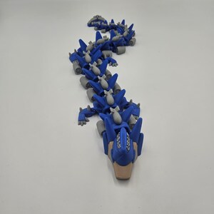 Pode incluir: Um brinquedo de drag&atilde;o articulado azul e cinza. O drag&atilde;o tem um corpo segmentado com placas azuis, detalhes cinzentos e pequenas bombas cinzentas. A cabe&ccedil;a &eacute; detalhada com um focinho bege e elementos azuis. O brinquedo est&aacute; sobre uma superf&iacute;cie branca.