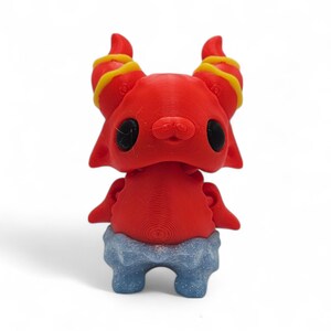 May include: A red figurine with large black eyes, small ears, and yellow-striped horns. The figurine has a blue and gray textured base, and a small, smiling face. The figure is a whimsical, cartoon-like character.