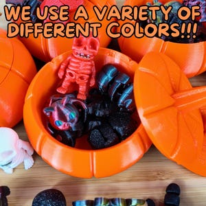 May include: A variety of small, colorful, 3D printed figurines are displayed inside orange pumpkin-shaped containers. The text "WE USE A VARIETY OF DIFFERENT COLORS!!!" is visible in the image.