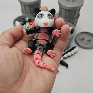 May include: A 3D-printed articulated opossum toy, held in a hand. The opossum has a white head, pink nose and tail, and black and gray body parts. Gray trash cans are in the background.