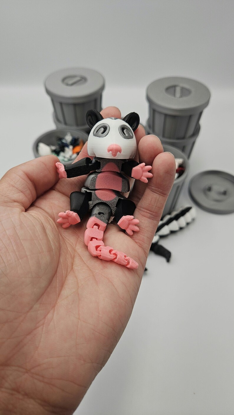 May include: A 3D-printed articulated opossum toy, held in a hand. The opossum has a white head, pink nose and tail, and black and gray body parts. Gray trash cans are in the background.