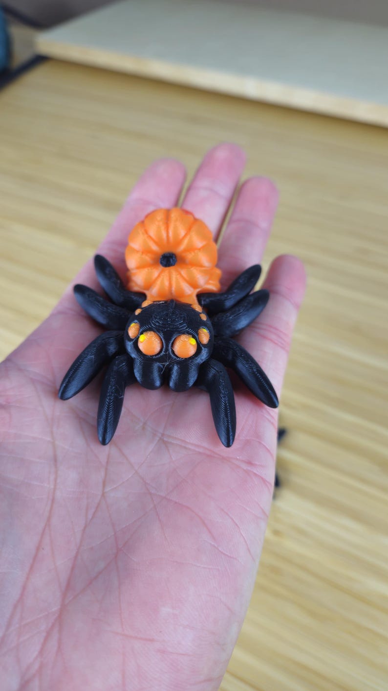 Puede incluir: Una ara&ntilde;a impresa en 3D de color negro y naranja con ojos grandes. La ara&ntilde;a tiene un cuerpo con forma de calabaza.