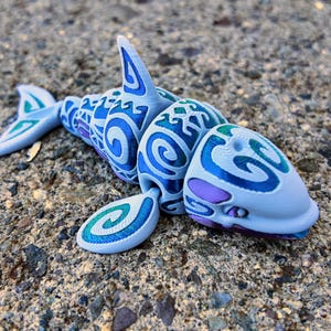 May include: A white, blue, and green plastic toy shark with a tribal pattern. The shark has a large, open mouth and a pink tongue.