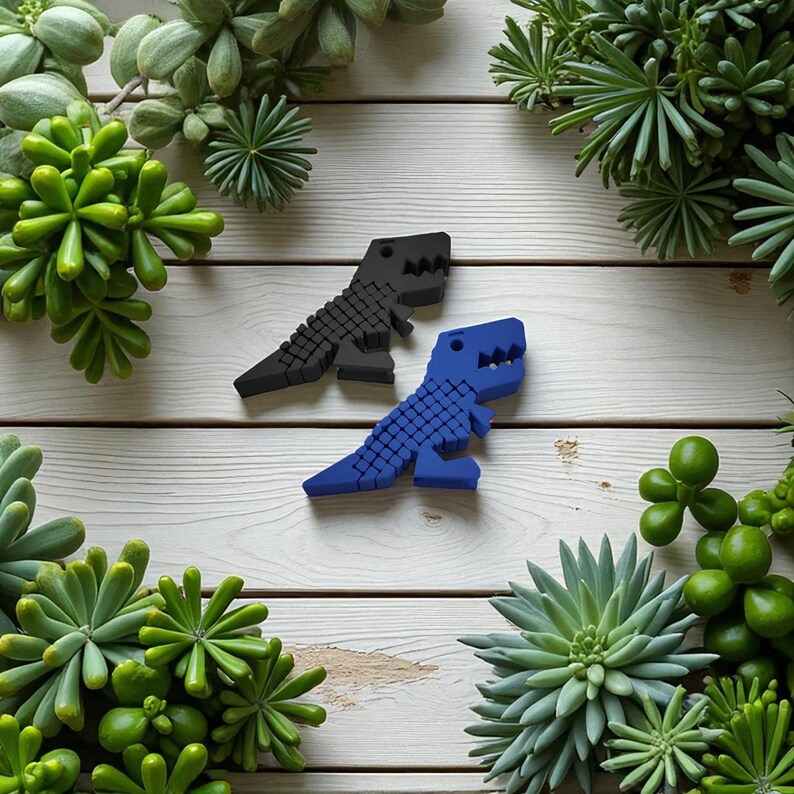 Puede incluir: Dos objetos con forma de dinosaurio, uno negro y otro azul, se muestran sobre una superficie de madera blanca. Los dinosaurios est&aacute;n hechos de peque&ntilde;os bloques cuadrados. Las suculentas verdes rodean a los dinosaurios, creando un tema decorativo natural.