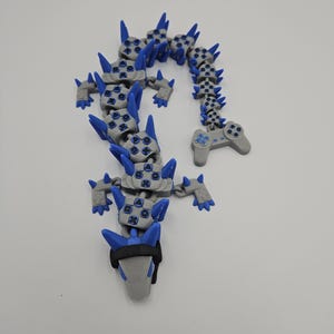 May include: A gray and blue articulated dragon toy with a video game controller design. The dragon features blue spikes and button details, with a black band around the head. The toy is designed to be flexible and poseable.