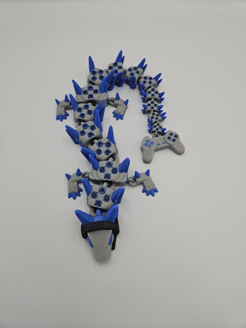 May include: A gray and blue articulated dragon toy with a video game controller design. The dragon features blue spikes and button details, with a black band around the head. The toy is designed to be flexible and poseable.