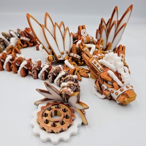 May include: A white and copper-coloured 3D printed dragon necklace with intricate details and a gear charm. The dragon has a long, slender body with wings and a tail. The gear charm is attached to the dragon's tail.