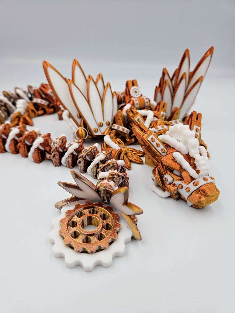 May include: A white and copper-coloured 3D printed dragon necklace with intricate details and a gear charm. The dragon has a long, slender body with wings and a tail. The gear charm is attached to the dragon's tail.