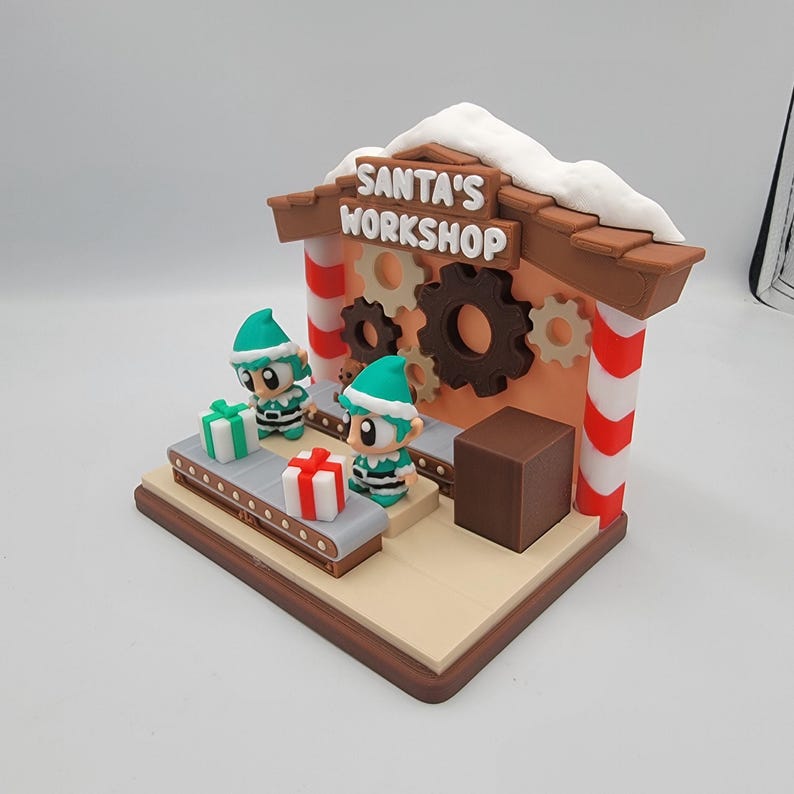 Może przedstawiać: Świąteczna dekoracja bożonarodzeniowa przedstawiająca Warsztat Świętego Mikołaja. Miniaturowa scena zawiera dwie figurki elf&oacute;w, przenośnik taśmowy z zapakowanymi prezentami oraz budynek z napisem "SANTA'S WORKSHOP". Budynek ma czerwono-białe kolumny w paski i ośnieżony dach.
