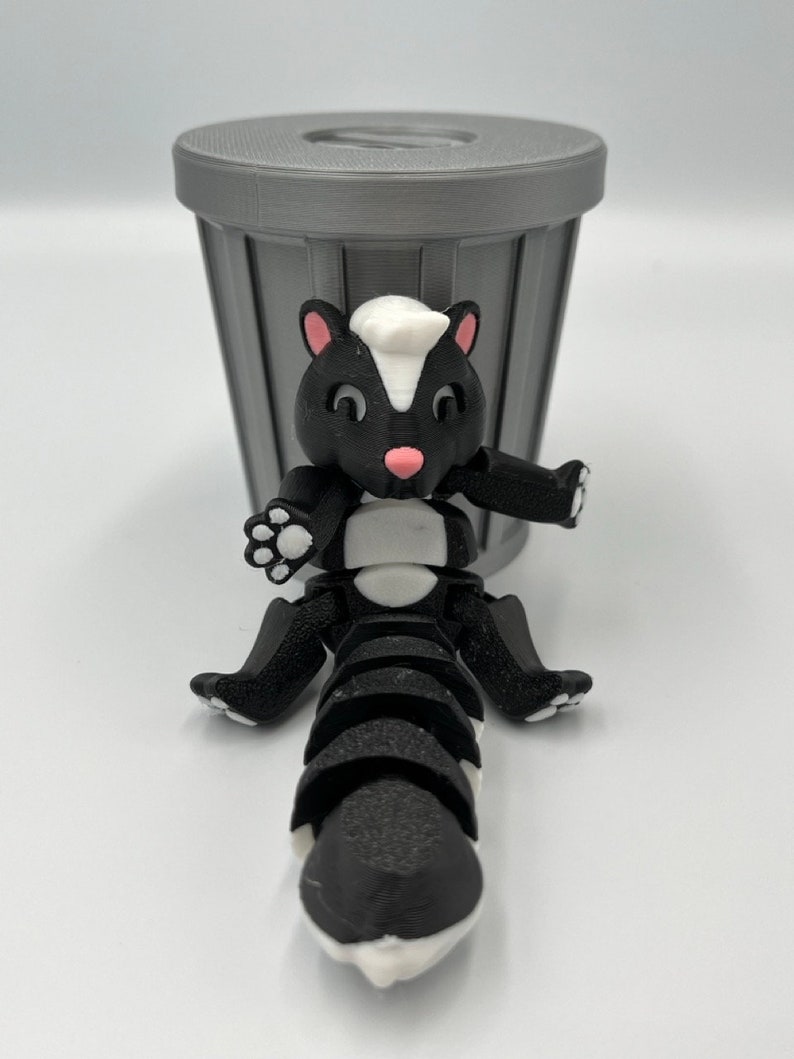 May include: A 3D-printed articulated skunk toy in black, white, and pink, posed in front of a gray trash can. The skunk has a white stripe down its back and a white patch on its head. The trash can is gray.