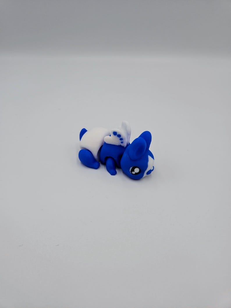K&ouml;nnte beinhalten: Ein kleines blaues und wei&szlig;es Plastikspielzeugkaninchen mit einem wei&szlig;en Ohr und einem wei&szlig;en Fleck auf der Nase. Das Kaninchen liegt auf der Seite mit dem Kopf nach rechts gedreht.