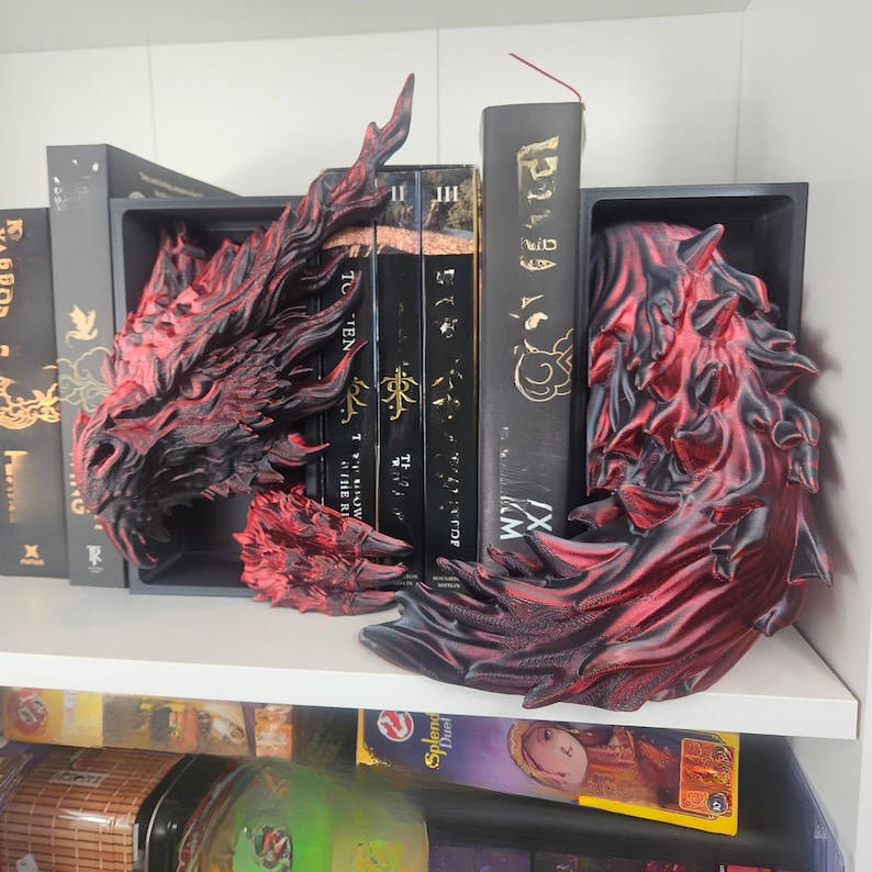 May include: A pair of dragon-themed bookends in black and red, with intricate detailing. The bookends are placed on a white shelf, holding up a row of black books. The dragon's head and tail are visible, adding a fantasy element to the bookshelf.