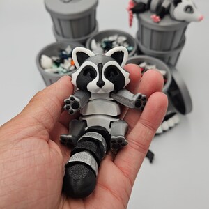 May include: A 3D-printed raccoon figurine with a black and white striped tail and a gray body. The raccoon is held in a hand, with several gray trash cans in the background, some with trash and a possum figurine.