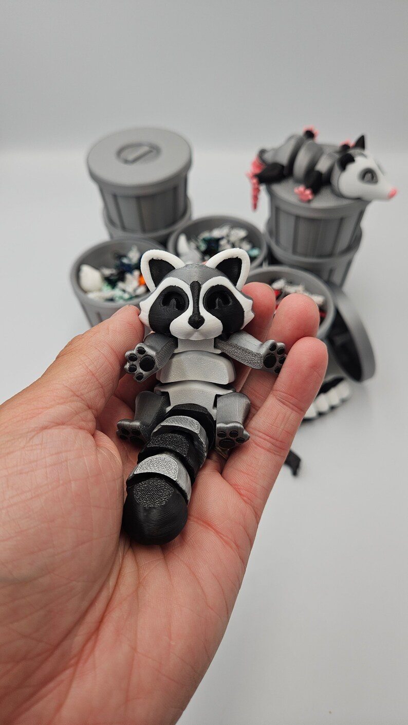 May include: A 3D-printed raccoon figurine with a black and white striped tail and a gray body. The raccoon is held in a hand, with several gray trash cans in the background, some with trash and a possum figurine.