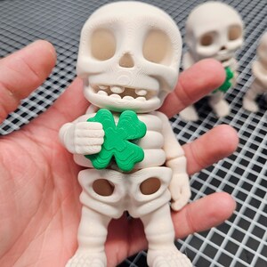 May include: A white skeleton figurine holding a green four-leaf clover. The figurine is standing on a hand.
