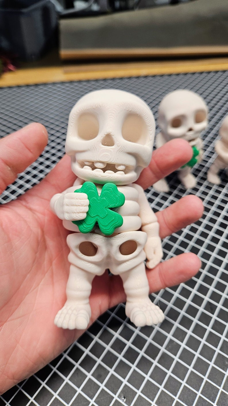 May include: A white skeleton figurine holding a green four-leaf clover. The figurine is standing on a hand.
