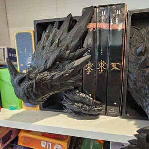 Op de afbeelding: Een zwarte drakenkop boekensteun, met gedetailleerde schubben en hoorns, staat naast een set boeken. De boekensteun is gemaakt van een textuurrijk materiaal en past in een boekenplank.