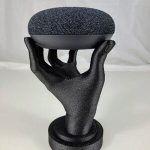 Op de afbeelding: Een zwarte Google Home slimme speaker staat op een zwarte handvormige standaard. De standaard heeft een ronde basis.