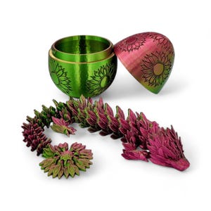 Puede incluir: Una figura de drag&oacute;n impresa en 3D de color verde y rosa con detalles de girasol. El drag&oacute;n est&aacute; enroscado y tiene una cola larga. El drag&oacute;n est&aacute; sentado junto a un huevo impreso en 3D de color verde y rosa con detalles de girasol. El huevo est&aacute; abierto y la mitad superior est&aacute; sentada junto al drag&oacute;n.