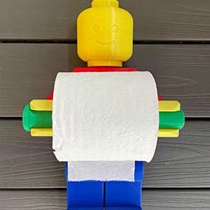 K&ouml;nnte beinhalten: Ein neuartiger Toilettenpapierhalter in Form eines gelben Lego-Figurenkopfes mit rotem Hals, gr&uuml;nen und gelben Armen und blauen Beinen. Der Halter ist f&uuml;r eine Rolle wei&szlig;es Toilettenpapier konzipiert.