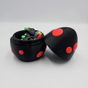May include: A black egg-shaped container with red polka dots. The container is open and reveals a small green, black, and blue dinosaur figurine inside.