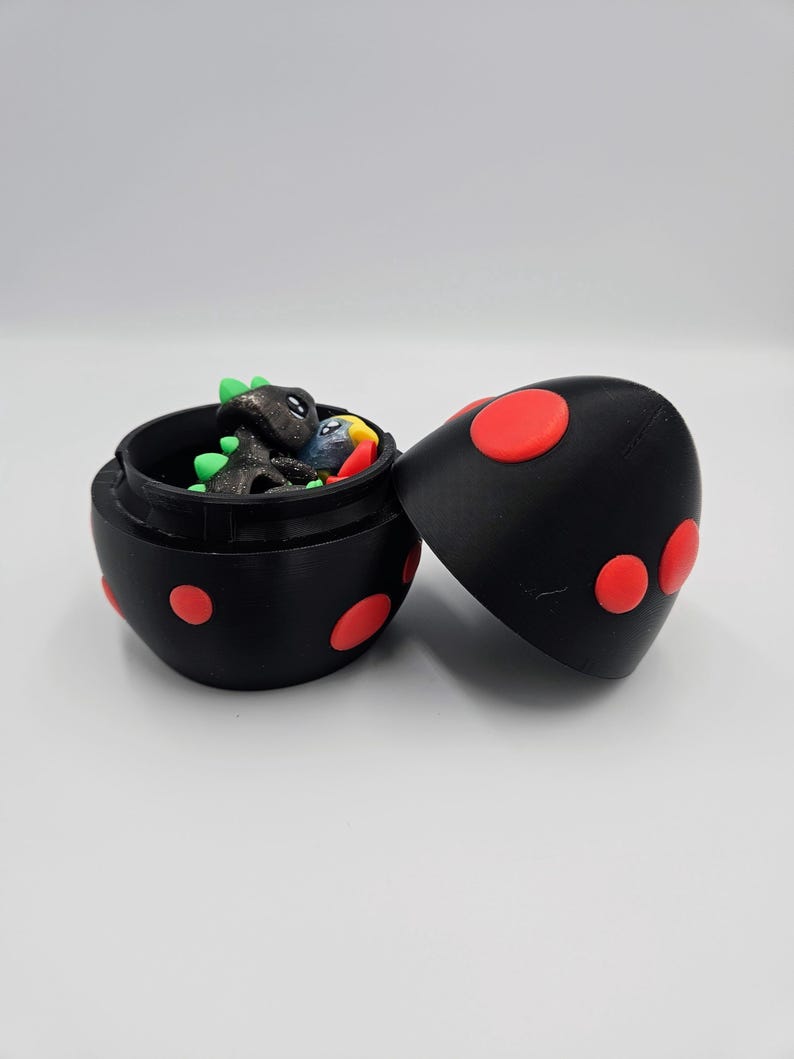May include: A black egg-shaped container with red polka dots. The container is open and reveals a small green, black, and blue dinosaur figurine inside.