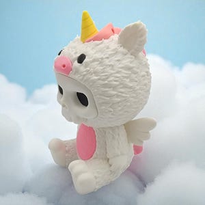 Puede incluir: Figura blanca de una calavera con un disfraz de unicornio, con cuerno amarillo, detalles rosas y peque&ntilde;as alas blancas. La figura est&aacute; sentada sobre una nube esponjosa contra un fondo de cielo azul.