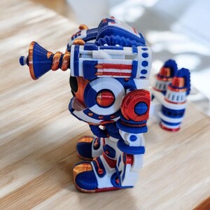 May include: A 3D printed robot toy in blue, white and red. The robot has a large head with a circular eye and a long, curved arm with a horn-like structure. The robot is standing on a wooden surface.