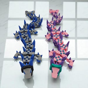 May include: Two articulated dragon sculptures. One is gray with blue accents, the other is pink with purple accents. Both have a segmented body, sharp spikes, and a controller-shaped head. The dragons are displayed on a white surface.