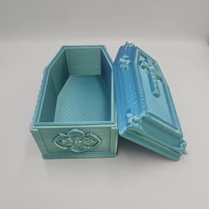 May include: An open, light blue coffin-shaped box with ornate detailing. The lid is ajar, revealing the interior. The box features a cross design on the lid and a floral emblem on the front. The box is made of a textured material.