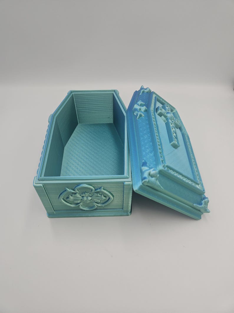 May include: An open, light blue coffin-shaped box with ornate detailing. The lid is ajar, revealing the interior. The box features a cross design on the lid and a floral emblem on the front. The box is made of a textured material.