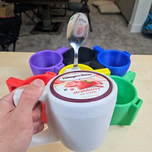 Pu&ograve; includere: Un supporto bianco a forma di tazza con manico, progettato per contenere una pinta di gelato. Il coperchio di un contenitore di gelato alla fragola &egrave; visibile, con un cucchiaio sopra. Sono visibili anche altri supporti colorati in rosso, verde, blu, giallo e viola.